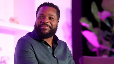 Variety via Getty Images Malcolm-Jamal Warner sitting onstage in 2023. He is holding a microphone with the Variety logo on it.