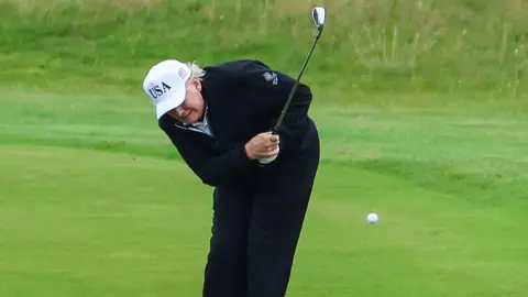 Reuters Donald Trump, wearing navy trousers and a navy jacket with a white "USA" cap, is doubled over as he strikes a golf ball.