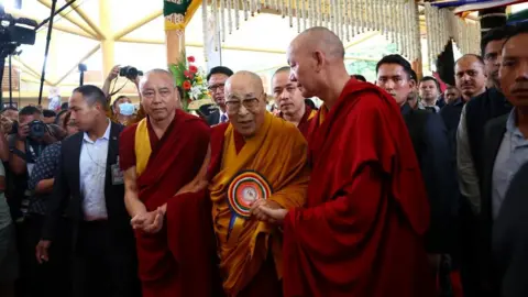 Reuters The bespectacled Dalai Lama cloaked in a red robe with a flowing orange wrap, arrives for his birthday celebrations flanked by two Buddhist's in red robes holding his hand. They are surrounded by suited men and a number of photographers.