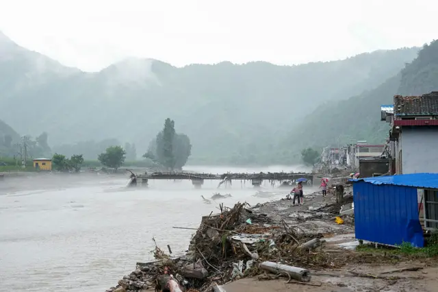 A river surrounded by hillsides with a destroyed bridge and debris on the banks