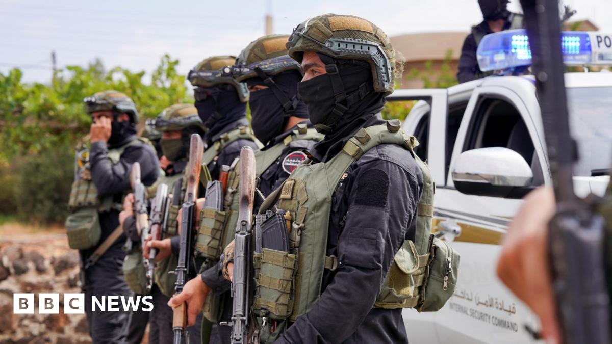 Arned Syrian security forces officers wearing green helmets and black masks, as well as protective vests, stand in a row in front of an emergency vehicle.