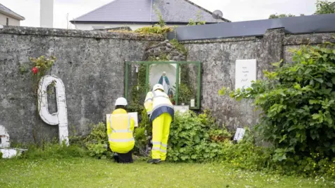PA Media Two workers in yellow high visibility clothing at a memorial left at the site of the  former Mother and Baby Institution in Tuam, County Galway.