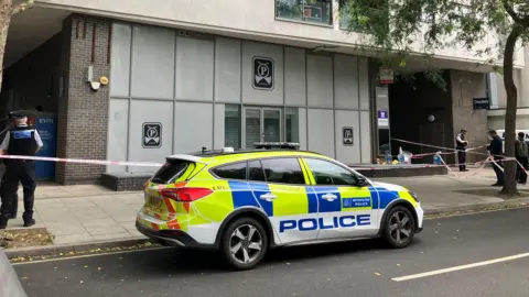 BBC A police car parked outside a grey building and several police officers nearby behind police tape 