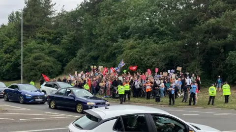 Alex Dunlop/BBC A large crowd of people standing by a junction. There is police around the group and protesters are holding flags.