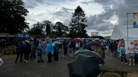 BBC Crowds of people at a festival. Many are holding umbrellas or wearing raincoats. Food and drink stalls are on the left and right, with portaloos in the background