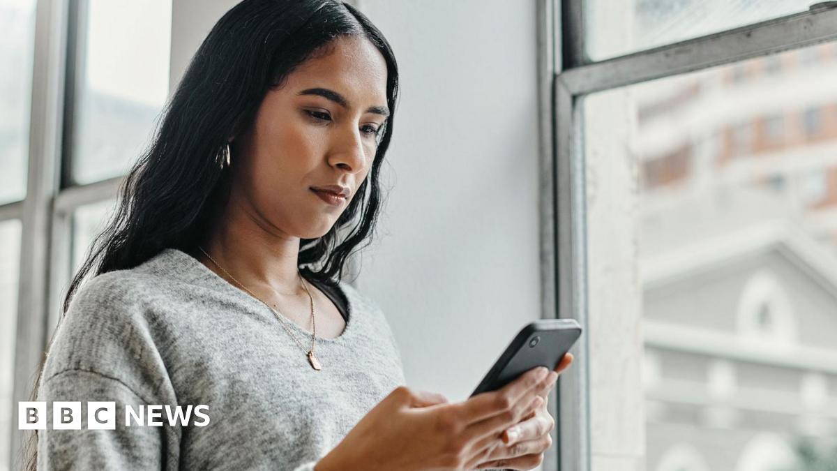 Young woman in grey pullover standing by a window and looking at her phone with a serious look on her face.