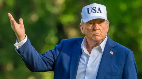Getty Images Donald Trump ,in a white USA cap and wearing a blue jacket with a US flag badge pinned to it, lift up one of his hands outside in the White House garden - 6 July 2025.
