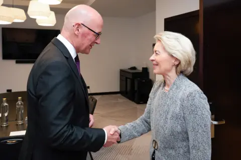European Commission's Audiovisual Service John Swinney, who has a bald head and is wearing a black suit jacket, shakes the hand of Ursula von der Leyen, who has short blonde hair and has grey suit jacket with a gold fastening.