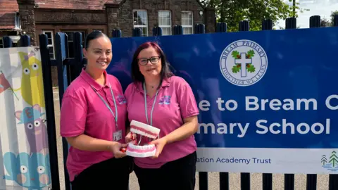 The image shows two women in pink tshirts holding a large fake mouth and teeth toy. They are standing outside in front of Bream CoE primary school. 
