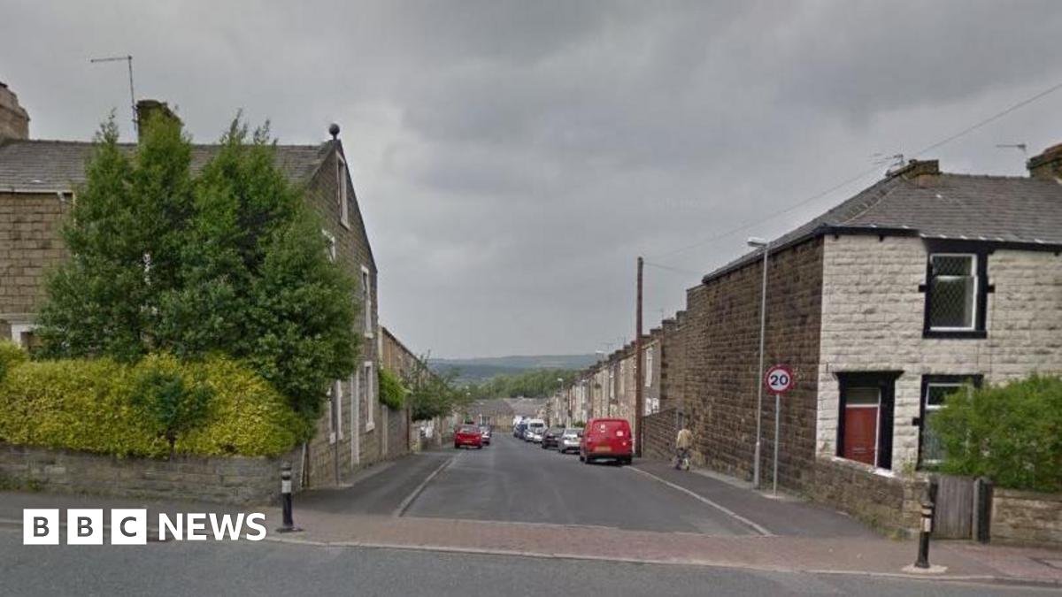 Google streetview looking down Craven Street, which is lined with dark and white brick terrace houses and parked cars. Pennine hills can be seen in the backdrop.