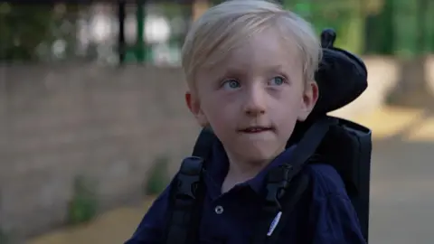 BBC A young boy with blond hair and light coloured eyes, wearing a dark blue top and sitting in a wheelchair.