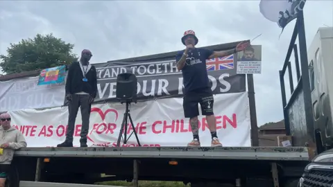 Richard Knights/BBC A man in black T-shirt and shorts and wearing a union jack bucket hat addresses a crowd from the back of a lorry. Another man stands beside him and looks on. Behind them are banners, one of which says "Unite as one for our children".