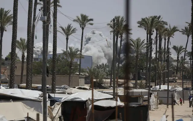 In distance smoke rises from Deir al-Balah in Gaza on 20 July and in foreground children run near tents and shelters