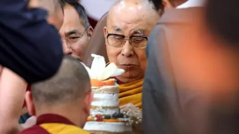 EPA/Shutterstock The bespectacled Dalai Lama cuts an iced white cake surrounded by people during his 90th birthday celebrations