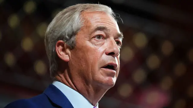 The side profile of Nigel Farage as he speaks at a conference, wearing a navy suit jacket and blue shirt.