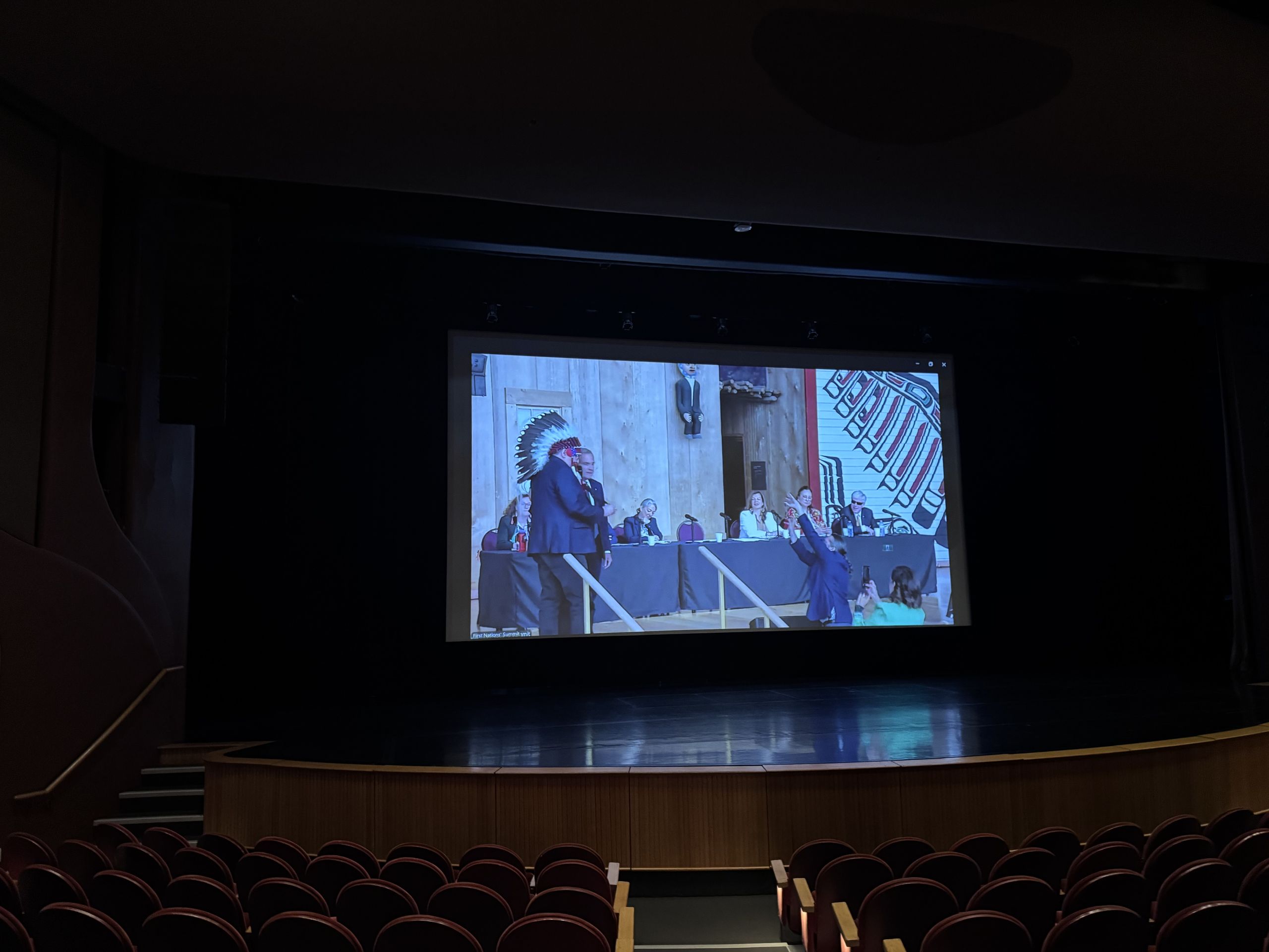 a screen showing a summit of First Nations leader at the front of a darkened auditorium
