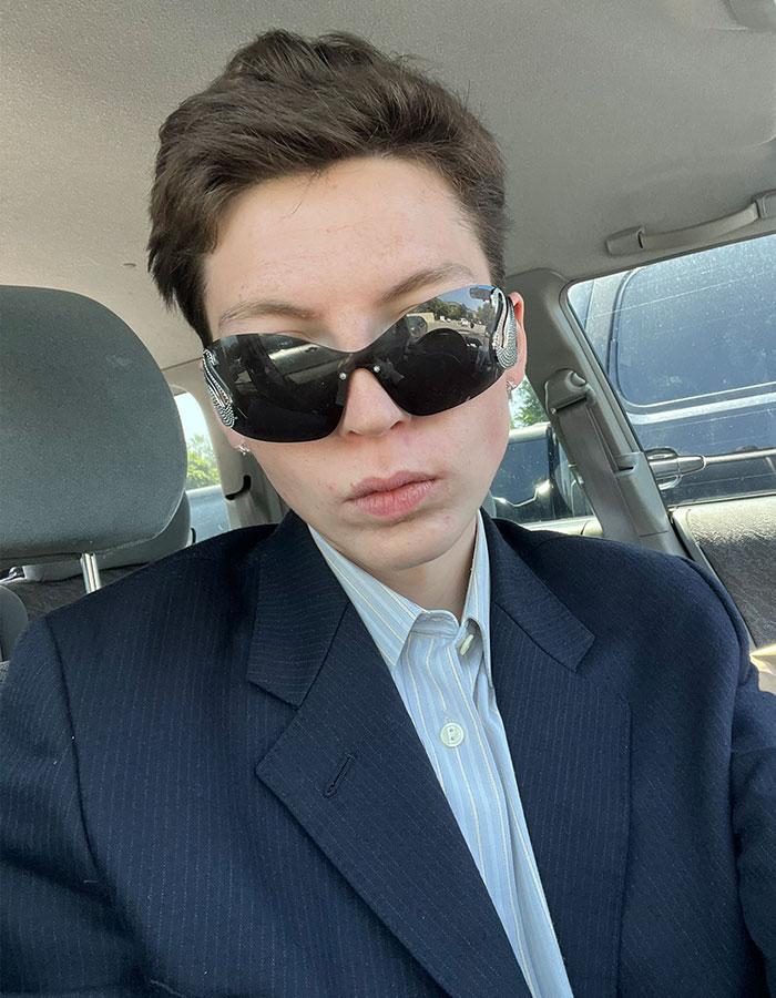 Person with short hair wearing dark sunglasses and a suit jacket sitting inside a car, related to model detransition story.