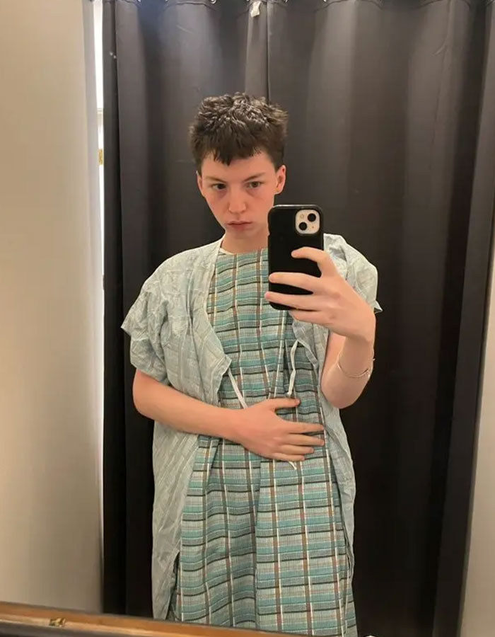 Young person in hospital gown taking a mirror selfie, representing model pushed to transition at 15 now detransitioning.