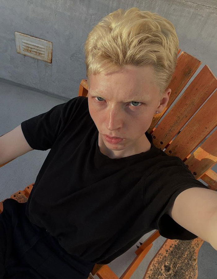 Model with short blonde hair sitting in an orange chair, reflecting on transitioning and detransitioning experience.