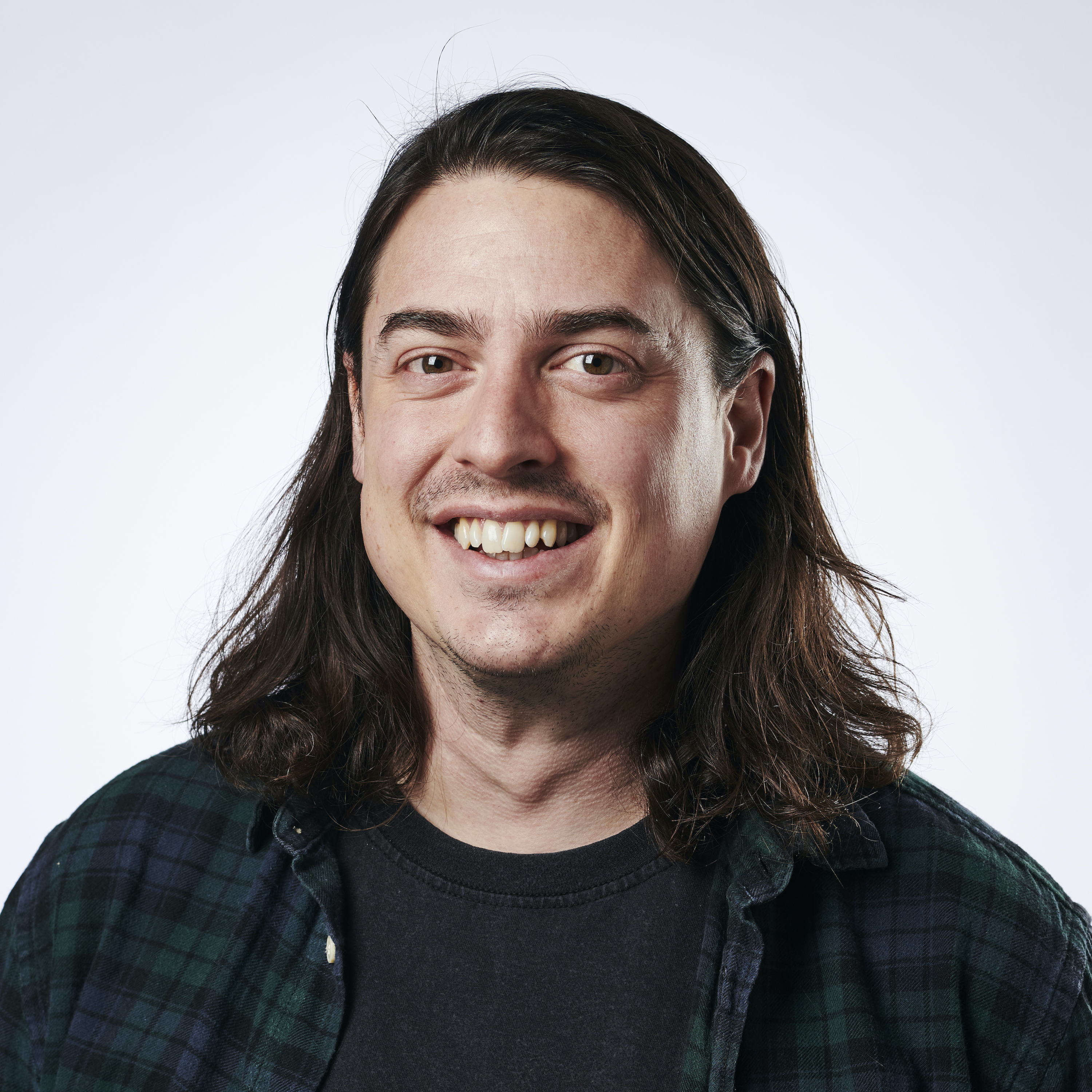 A photo of What Hi-Fi? journalist Tom Parsons. He is wearing an unbuttoned shirt over a t-shirt, has shoulder-length dark hair and is smiling at the camera. 