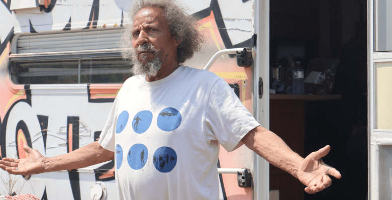 A senior Black man, with both arms spread, looks on with a disappointed expression, as if he has been wronged. Behind him is a trailer with an open door and his belongings on a table.