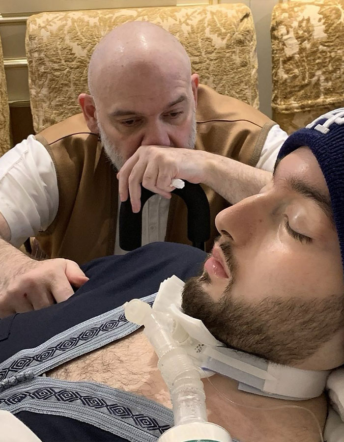 Man looking sorrowfully at a young man in a coma with a breathing tube, representing the sleeping prince of Saudi Arabia.