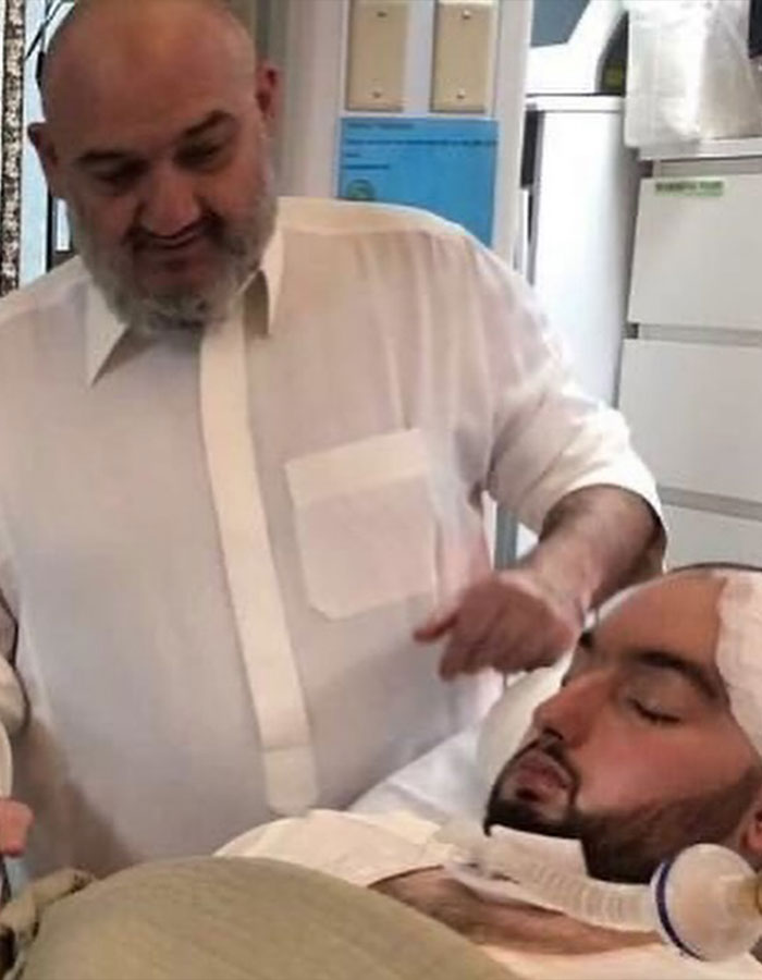 Man wearing a white shirt standing next to a bed with the Sleeping Prince of Saudi Arabia in a coma.