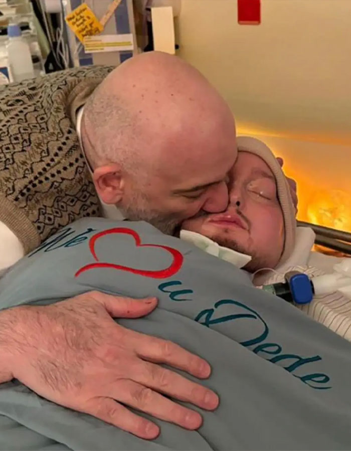 Man kissing the sleeping prince of Saudi Arabia, who is in a hospital bed covered with a blanket showing love and care.