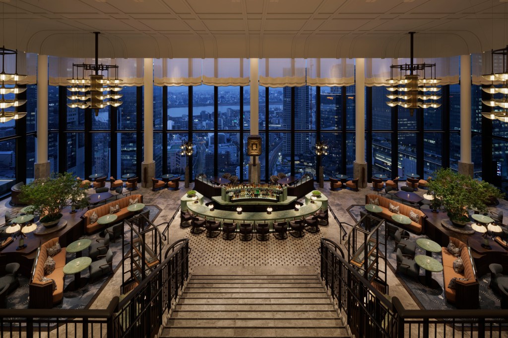 Waldorf Astoria Osaka - Peacock Alley at Night - floor to ceiling windows overlooking the city skyline at night, staircase leading down to a central bar surrounded by a variety of table and seating on either side