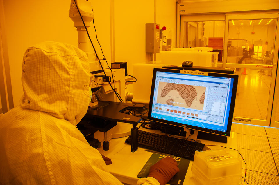 In a lab with orange lighting, a scientist wearing a full suit of protective gear sits at a table with a microscope and looks at images on a screen.