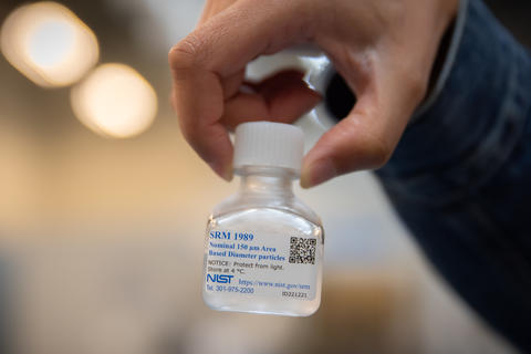 A hand holds a small, clear bottle full of a liquid solution with a label that says SRM 1989.