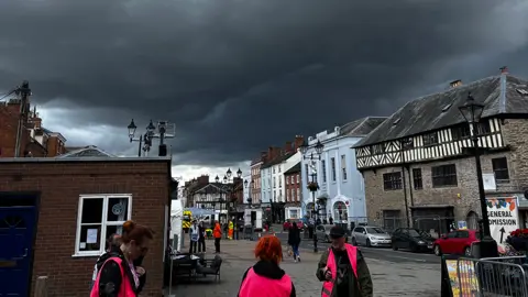 Dark clouds over Ludlow's market square. Three event stewards are in the foreground in pink high-vis jackets. Buildings, some of them black and white timber framed, are across
 the street