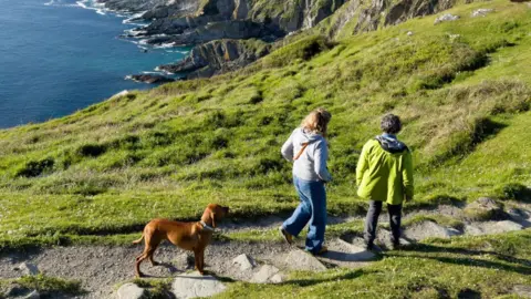 Getty Images Two people are walking along a grassy path accompanied by a brown dog. The path winds toward dramatic, rocky cliffs that drop into the sea.