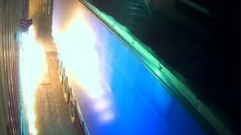 Metropolitan Police A security camera's view captured the fire at the warehouse as it licks towards the lorry in Leyton, east London