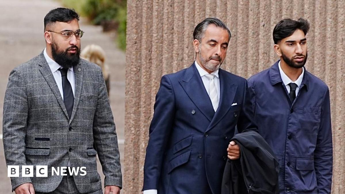 A young man with a dark beard wears a white shirt and a black tie and jacket and walks along a street.