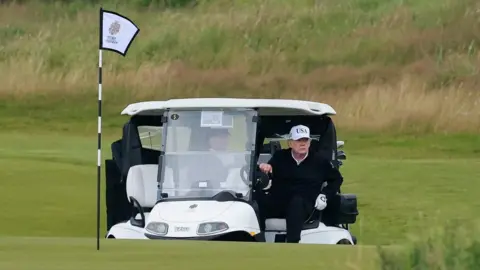 Reuters President Trump, wearing black clothes and a white baseball cap, gets out of a white golf buggy on the Turnberry course. There is a flag in the foreground.