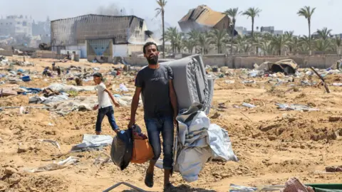 Reuters A displaced Palestinian man carries his possessions following an Israeli military ground operation in Deir al-Balah, central Gaza (22 July 2025)