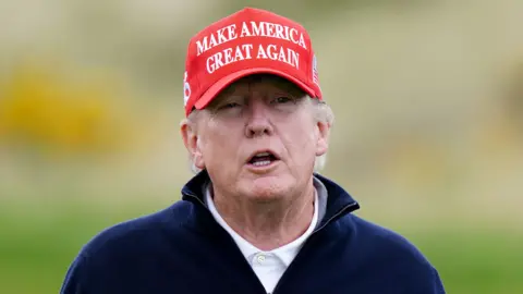 PA Media Donald Trump looking slightly off camera. He is only visible from the shoulders up. He has a red cap on his head with the words: 'Make America Great Again' in white writing on the front. He is wearing a white polo shirt under a dark blue zip-up jumper.