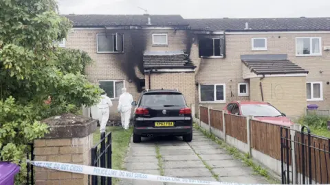 Jonny Humphries/BBC Two terraced houses with visible smoke damage on the brickwork and windows. Two forensic investigators in white overalls approach the front door of the house on the left, which is cordoned off with blue and white police tape. 