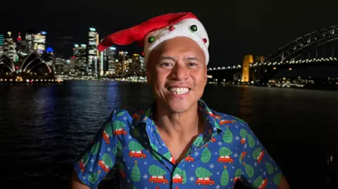 James Chater/BBC News A man in a Christmas themed shirt and wearing a Santa hat smiles at the camera with Sydney harbour in the background