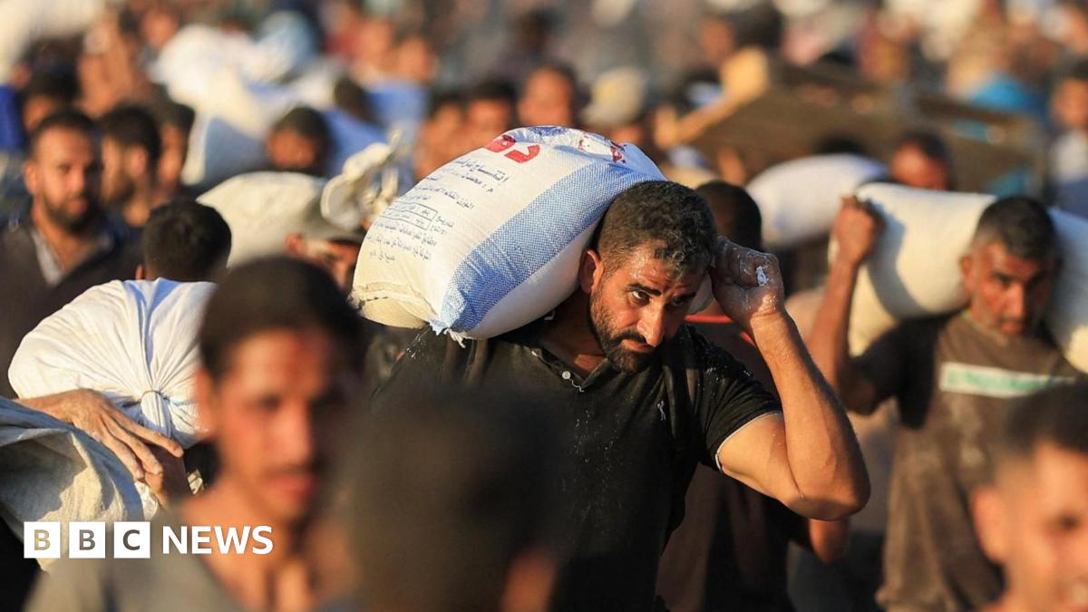 A man holding a large bag of aid over his shoulder is in focus in the foreground. He is one of several men carrying aid in the photo, with the rest blurred into the background.
