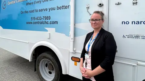 Canadian Press Catalina Friesen, a personal support worker and Low German-speaking liaison, stands in front of a bus outfitted as a mobile walk-in clinic, in St. Thomas, Ontario. She has a slight smile on her face and is wearing a dark blazer and a white T-shirt