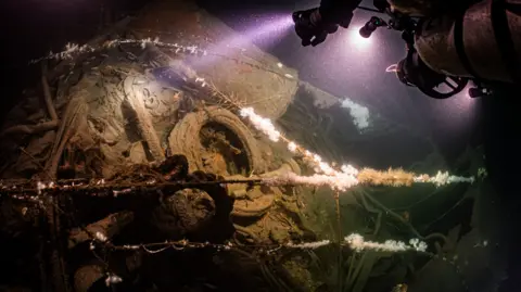 Steffen Scholz Divers shine a light at the wreckage of a royal navy warship, revealing rigging and warped metal covered in moss.