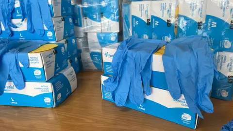 Boxes and blue and white boxes of PPE gloves sit on a table, with several pairs of blue plastic gloves draped across the front. 