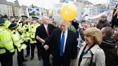 Getty Images A large group of protestors with placards outside the Scottish Parliament. Donald Trump, in dark coat with white shirt and light blue tie, is moving through them. A man is running a large orange balloon on Trump's head.