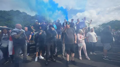PA Media A group of mostly male protesters, some standing on the back of flatbed truck with a white cab. Some are wearing caps or face coverings, while others point their mobile phone cameras towards the photographer. The air is smoky from flares and plumes of blue smoke are rising up from the crowd.