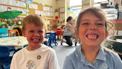 BBC The image shows two Year One pupils sat with big teethy grins showing their teeth covered in toothpaste. The picture is taken in a class room and the children have both got stickers on their school uniforms. 