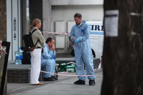 PA Media Two police officers in blue forensic overalls behind police tape speak to a woman in white trousers who is wearing a black rucksack and folding her arms