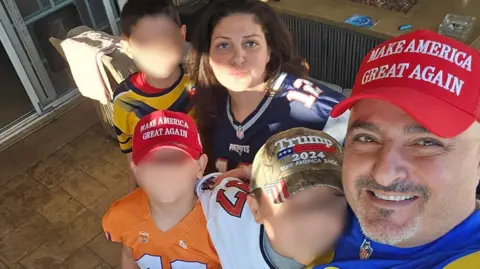 Arthur Sahakyan A family of six pose for a selfie taken by the dad Arthur who is wearing a red Make America Great Again hat. Two of the young boys wear MAGA/Trump hats as well. The mom and dad and all four children are wearing football jerseys from different teams. 