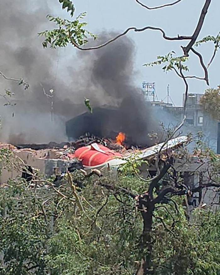 Wreckage of Air India plane crash with smoke and flames rising above building, showing aftermath of the disaster.